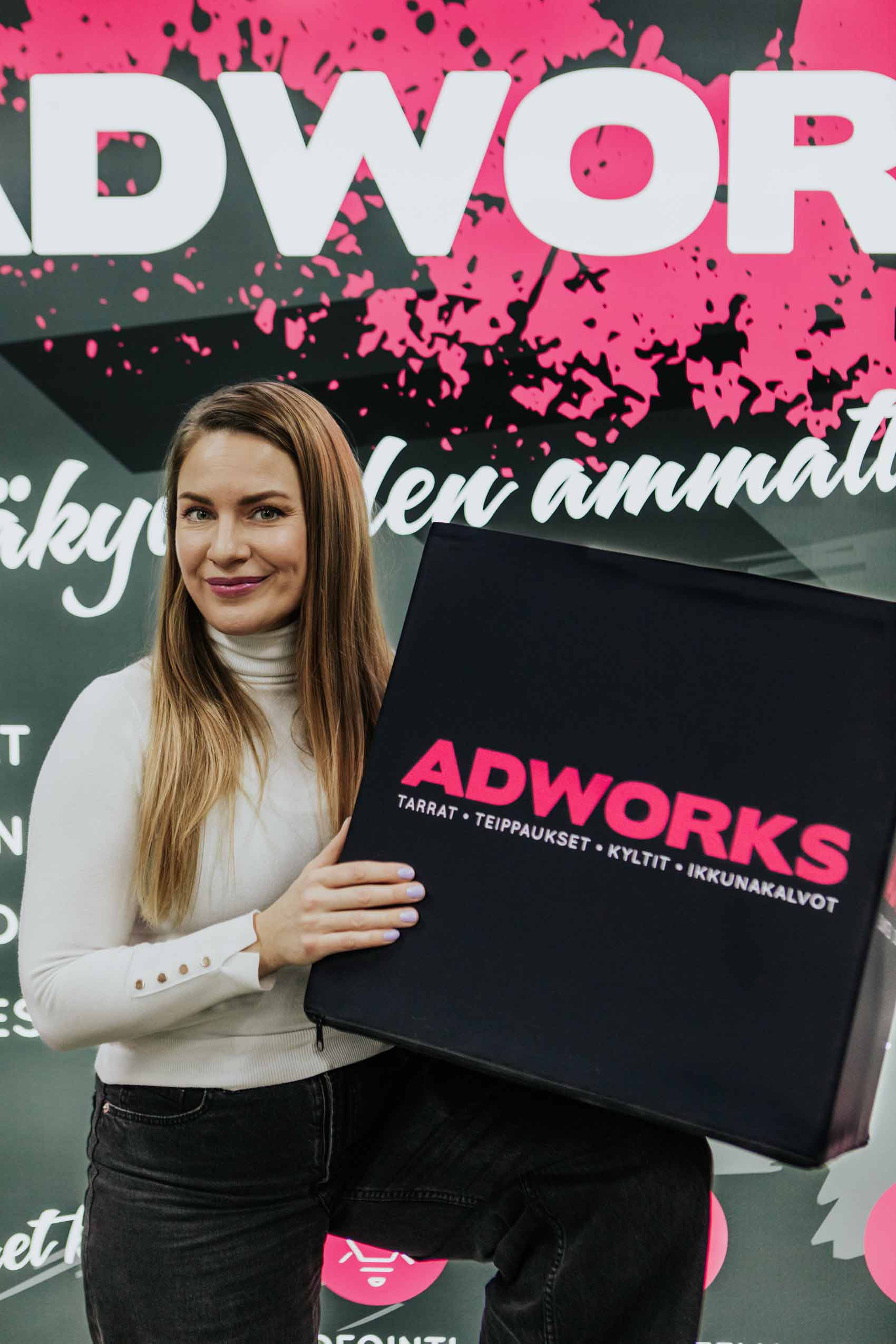 Nonna Achrén hymyilee ja katsoo kameraan pidellen tumman harmaata kuutiota jossa on magenta Adworksin logo. Taustalla on harmaata magentaa.
